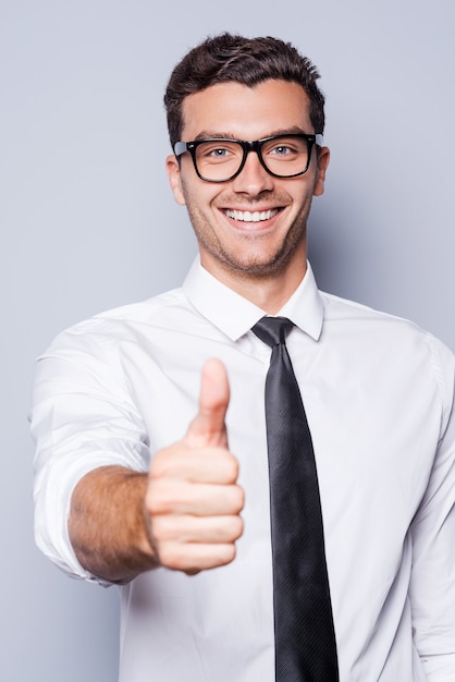 Buon lavoro! Felice giovane uomo in camicia e cravatta che mostra il pollice in su e sorride mentre si trova in piedi su sfondo grigio