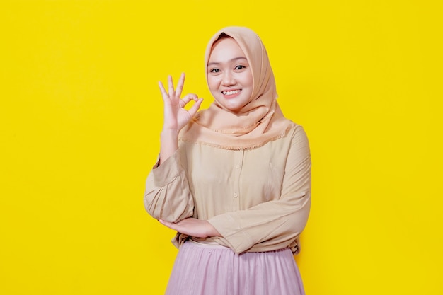 Buon lavoro orgogliosa felice giovane donna asiatica che indossa l'hijab mostra un gesto ok che fa l'occhiolino gioioso isolato su sfondo giallo