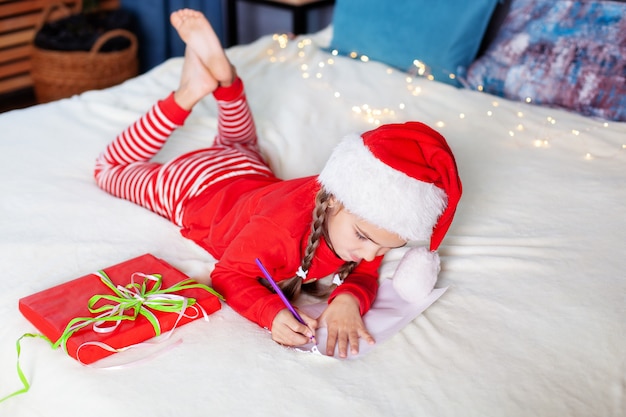 Buon Natale. Capodanno 2020! bambina in pigiama rosso e cappello di Babbo Natale scrive lettera a Babbo Natale in camera da letto