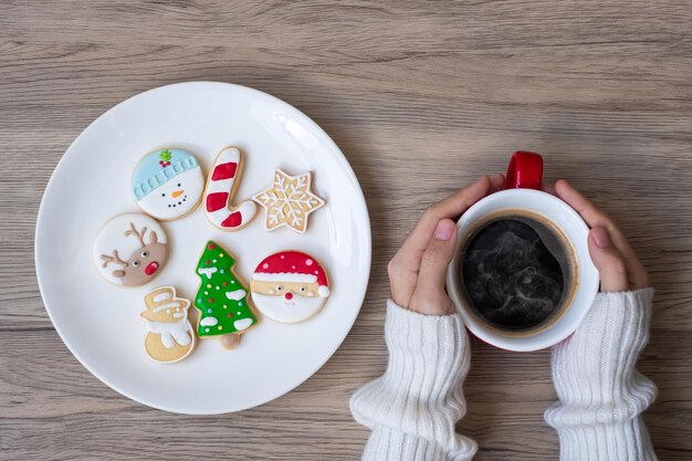 Buon Natale con la mano della donna che tiene la tazza di caffè e il biscotto fatto in casa sul tavolo. Vigilia di Natale, festa, vacanza e concetto di felice anno nuovo
