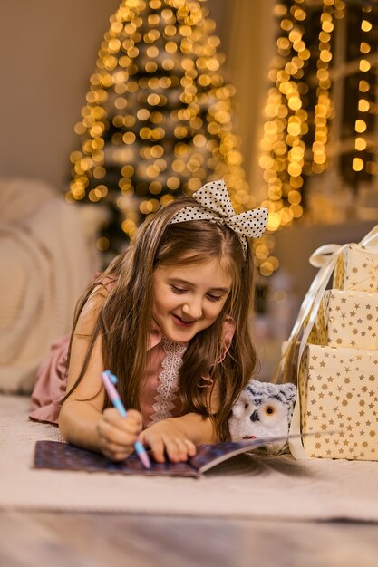 Buon Natale e Buone Feste. La bambina carina scrive la lettera a Babbo Natale vicino all'albero di Natale al chiuso.