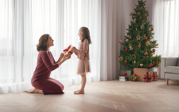 Buon Natale e buone feste. Madre allegra e sua figlia carina che si scambiano regali. Mamma e bambino che si divertono vicino all'albero all'interno. Famiglia amorevole con regali in camera.