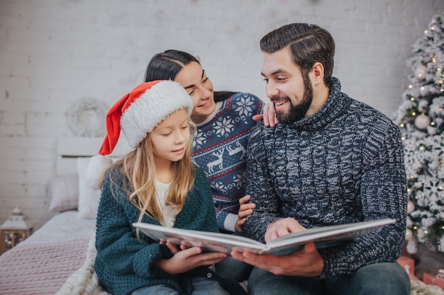 Buon Natale e buone feste Mamma allegra, papà e sua figlia carina che leggono un libro. Genitore e bambino divertirsi vicino all'albero di Natale in casa. Natale del mattino. Ritratto di famiglia da vicino