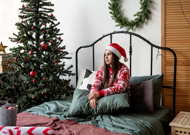 Buon Natale e Felice Anno nuovo. Felice giovane donna in maglione rosso e cappello da Babbo Natale seduta sul letto che celebra il natale