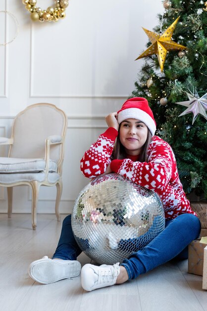 Buon Natale e Felice Anno nuovo. Giovane donna castana in cappello di santa che tiene palla da discoteca specchio seduto vicino all'albero di Natale