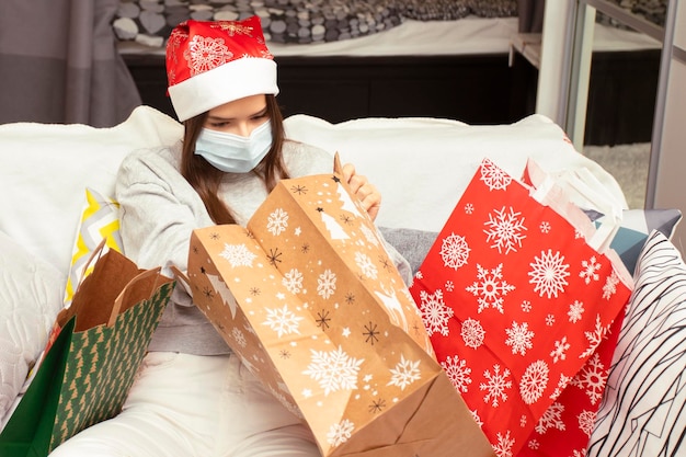 Buon Natale e felice anno nuovo La giovane donna in quarantena che indossa una maschera medica a casa guarda i regali in borse con motivi invernali durante l'epidemia di coronavirus