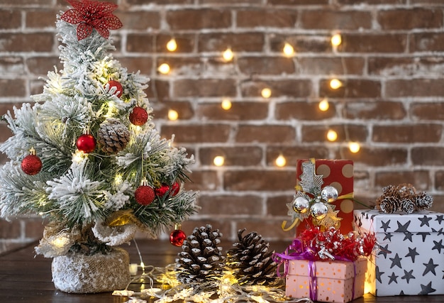 Buon Natale e felice anno nuovo, piccolo albero di Natale sul tavolo di legno con palline rosse e luci. Regali di Natale con decorazioni. Sfondo muro di mattoni
