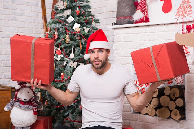 Buon Natale e Felice Anno nuovo. Uomo sorpreso con scatole rosse all'albero di Natale al camino. Concetto di giorno di Santo Stefano. Macho in santa cappello tenere regali avvolti. Preparazione e celebrazione delle vacanze.