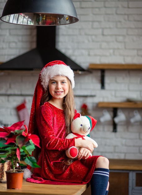 Buon Natale. La bambina sveglia in cappello di Santa sta tenendo un orsacchiotto che si siede nella cucina, aspettando la vacanza.
