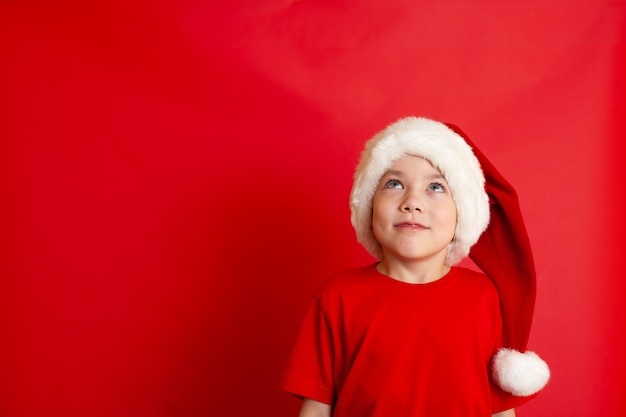 Buon Natale. Ritratto di un simpatico ragazzo allegro con un berretto natalizio in una maglietta rossa su sfondo rosso. Un posto per il testo. Foto di alta qualità