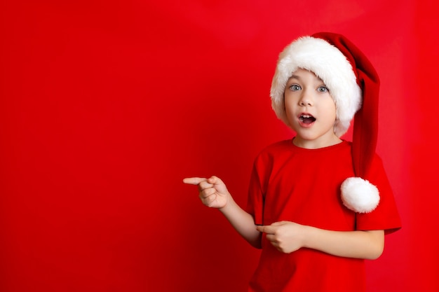 Buon Natale. Ritratto di un simpatico ragazzo allegro con un berretto natalizio in una maglietta rossa su sfondo rosso. Un posto per il testo. Foto di alta qualità