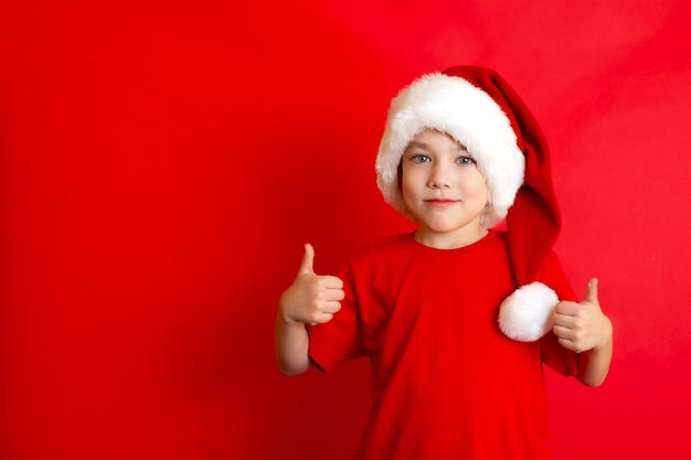 Buon Natale. Ritratto di un simpatico ragazzo allegro con un berretto natalizio in una maglietta rossa su sfondo rosso. Un posto per il testo. Foto di alta qualità