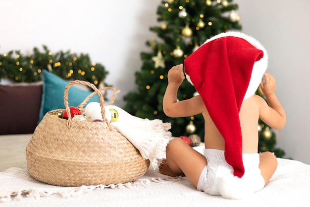 Buon Natale Un bambino con un pannolino e un cappello di Babbo Natale si siede su un letto in un'accogliente camera da letto decorata per l'albero di Natale natalizio Stile di vita