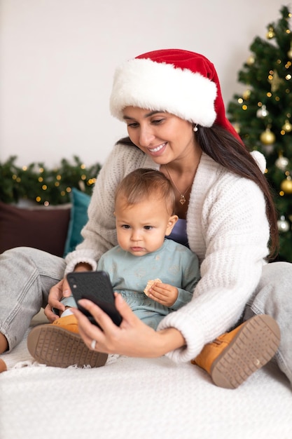 Buon Natale Una bella madre e una figlia piccola chiamano i loro parenti tramite collegamento video o si fanno un selfie su uno smartphone Accogliente camera da letto arredata Stile di vita