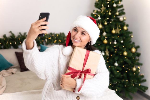 Buon Natale Una bellissima giovane donna con il cappello di Babbo Natale fa una videochiamata alla sua famiglia sdraiata in camera da letto con un albero di Natale decorato e luci sullo sfondo Stile di vita