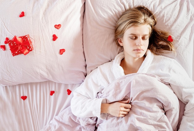 Buon San Valentino. giovane donna bionda che dorme a letto con regalo di festa e coriandoli rossi