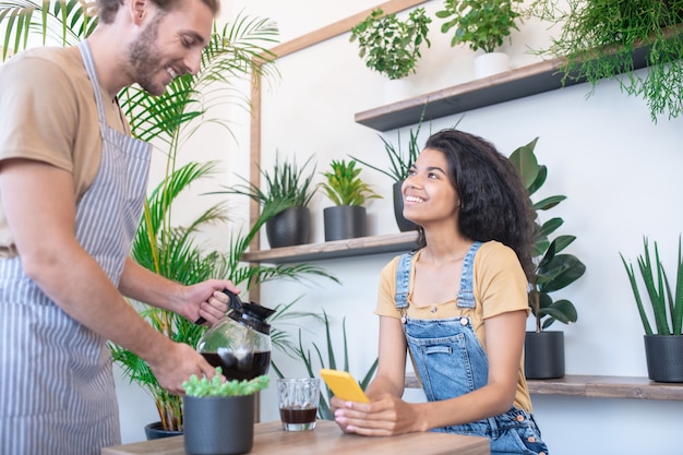 Buon servizio, caffetteria. Giovane uomo adulto amichevole in grembiule a strisce versando il caffè dalla caffettiera alla donna sorridente che si siede nella caffetteria