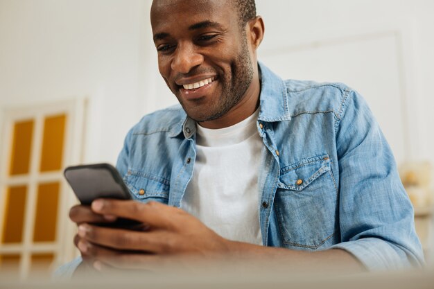 Buon umore. Bello giovane afroamericano sveglio che sorride e che scrive un messaggio sul suo telefono e che guarda lo schermo