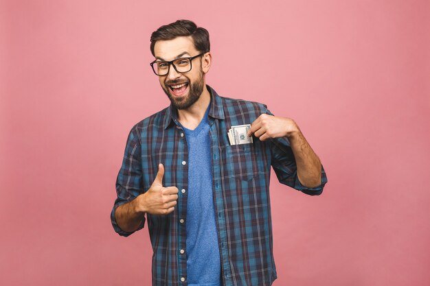 Buon vincitore! Giovane uomo ricco in banconote in dollari dei soldi della tenuta della camicia a quadretti nella tasca con la sorpresa isolata sopra fondo rosa.