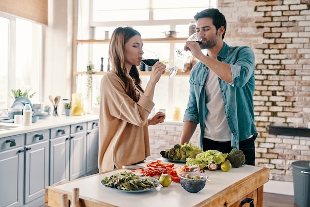 Buon vino in buona compagnia. Bella giovane coppia che cucina la cena e beve vino stando in piedi in cucina a casa