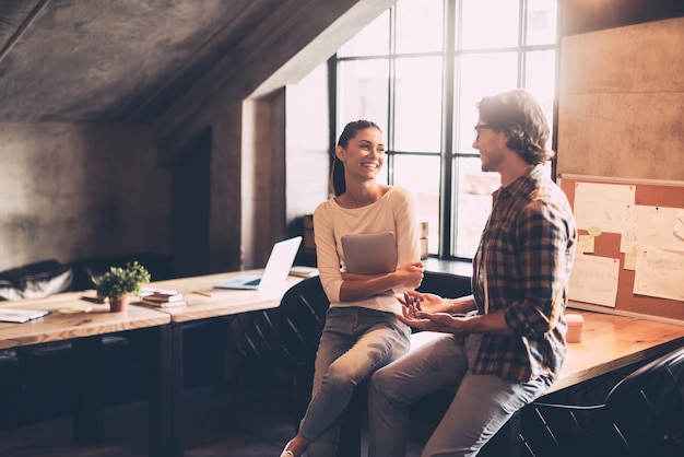 Buona chiacchierata con il collega. Allegro giovane e donna che discutono di qualcosa e sorridono mentre entrambi si appoggiano alla scrivania di legno nell'ufficio creativo creative