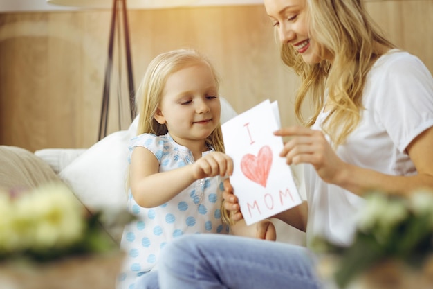 Buona festa della mamma in appartamento soleggiato! La figlia del bambino si congratula con la mamma e le dà la cartolina con il disegno del cuore. Concetto di famiglia.