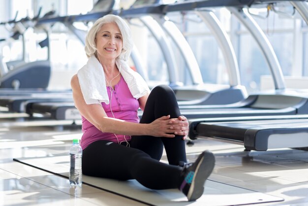 Buona giornata. Bella piacevole donna senior sorridente e utilizzando le cuffie mentre è seduto sul carped in una palestra.