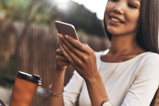 Buona giornata di lavoro. Attraente giovane donna che tiene in mano uno smartphone e sorride mentre è seduta al ristorante all'aperto