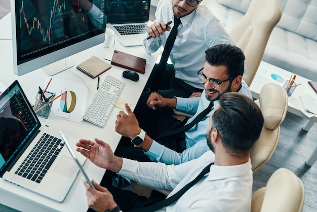 Buona giornata lavorativa. Vista dall'alto di giovani uomini moderni in abiti da cerimonia che analizzano i dati del mercato azionario e sorridono mentre lavorano in ufficio