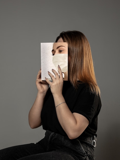 Buona giornata mondiale del libro 2020, sii al sicuro e leggi per diventare qualcun altro - donna che copre il viso con un libro in maschera mentre legge su sfondo grigio studio. Celebrazione, educazione, arte, concetto di protezione.