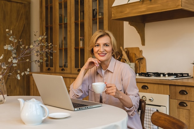 Buona mattinata. Allegra donna anziana sorridente in piedi in cucina e usando il suo computer portatile mentre riposa dopo colazione, libero professionista che lavora a casa.