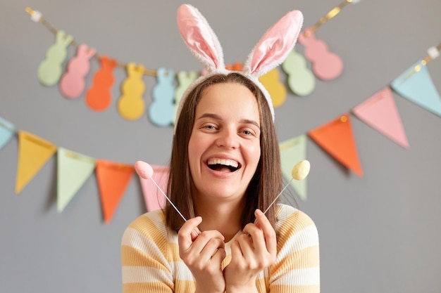 Buona Pasqua Affascinante donna sorridente con i capelli scuri che indossa orecchie di coniglio che tiene una deliziosa torta di Pasqua si apre ridendo guardando la fotocamera isolata su sfondo grigio decorato