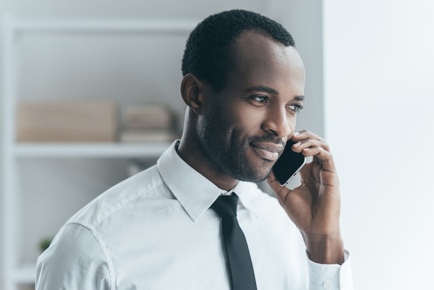 Buone chiacchiere d'affari. Bel giovane africano in abiti da cerimonia che parla al telefono e sorride mentre si trova in un ufficio creativo