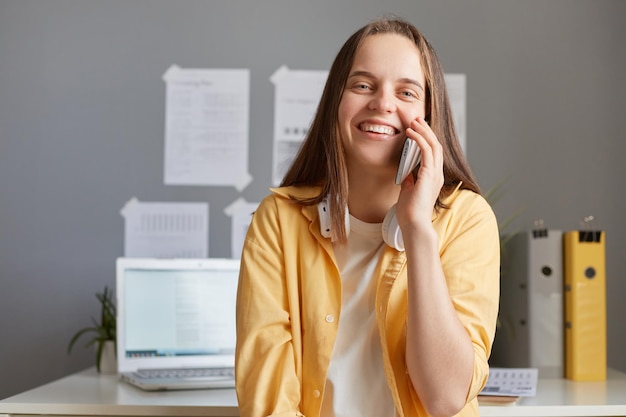 Buone conversazioni d'affari Allegra giovane bella donna hipster che indossa una giacca gialla che parla al cellulare mentre è seduta al suo posto di lavoro guardando la fotocamera con un sorriso