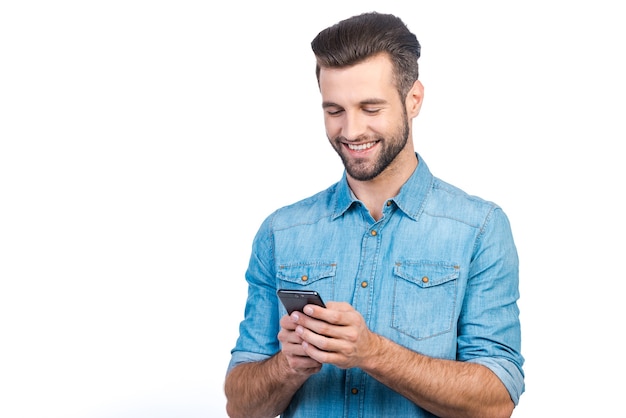 Buone notizie dall'amico. Fiducioso giovane bell'uomo in camicia di jeans che tiene in mano uno smartphone e lo guarda con un sorriso mentre è in piedi su sfondo bianco white