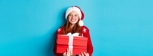 Buone vacanze e concetto di natale Allegra ragazza adolescente con i capelli rossi che indossa il cappello di Babbo Natale e tiene