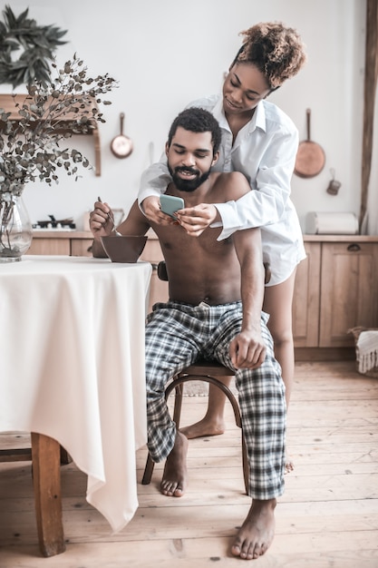 Buongiorno. Giovane marito barbuto dalla carnagione scura a torso nudo facendo colazione a tavola e la moglie che mostra lo schermo dello smartphone mattina in cucina