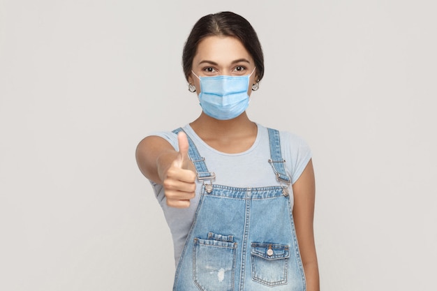 È buono, mi piace. Ritratto di giovane donna bruna soddisfatta con maschera medica chirurgica in tuta di jeans in piedi, pollice in alto e guardando la fotocamera. studio al coperto colpo isolato su sfondo grigio.