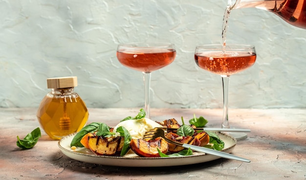 Burrata e pesche grigliate con basilico e pinoli Due bicchieri di vino rosato su sfondo chiaro Delizioso antipasto posto per testo vista dall'alto