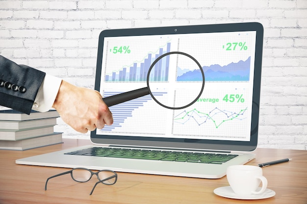 Businessman holding magnifier over laptop screen with business chart on wooden desktop with coffee cup glasses pen and books White brick wall background 3D Rendering