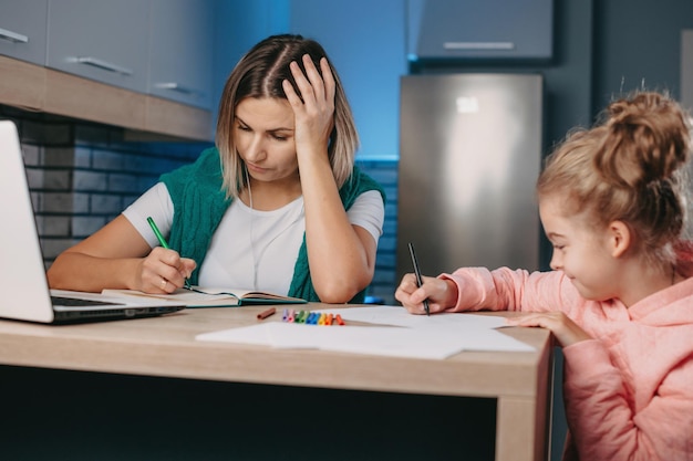 Bussineswoman che lavora al laptop da casa vicino a sua figlia che sta disegnando online l'attività di insegnamento woma...