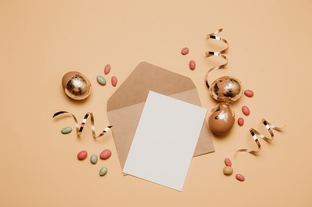 Busta in bianco del mestiere con un foglio di carta colto su una priorità bassa beige. Uova di Pasqua dorate con la caramella dei fagioli di gelatina, vista superiore.