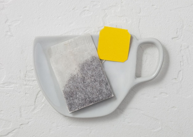 Bustina di tè al tè nero con etichetta gialla su piatto in ceramica a forma di tazza su sfondo chiaro