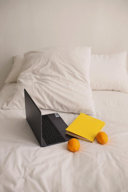 C'è un elegante laptop su un letto bianco accanto ad esso c'è un diario giallo e due mandarini maturi