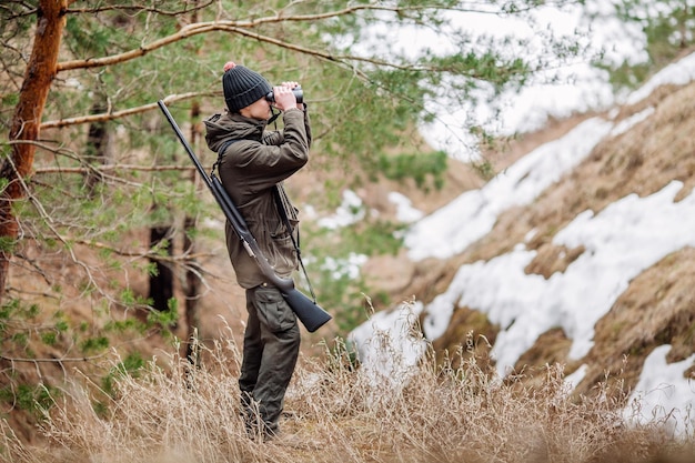 Cacciatore maschio con il binocolo pronto a cacciare tenendo la pistola e camminare nella foresta