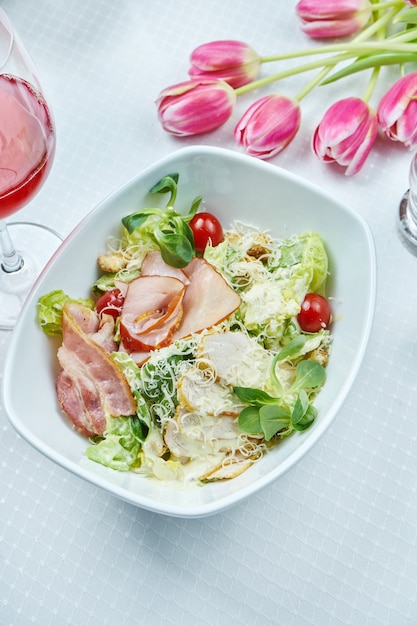 Caesar Salad con crostini di pane, parmigiano, pancetta, pollo, uovo in una ciotola bianca su bianco.