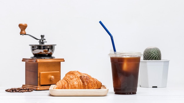 Caffè americano di ghiaccio in una tazza con croissant sul piatto di legno e macinacaffè pronto per il pasto della colazione