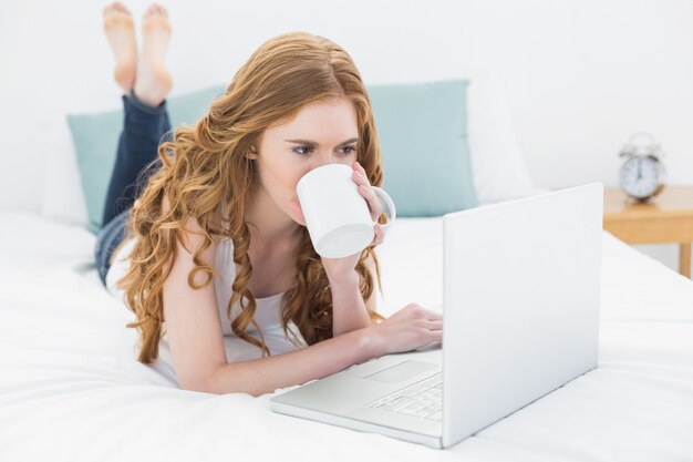 Caffè bevente biondo casuale mentre usando il computer portatile a letto