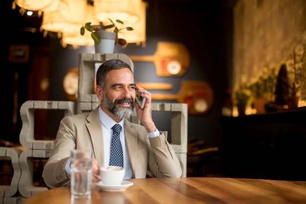 Caffè bevente dell'uomo d'affari maturo bello in caffè e nel telefono cellulare usando