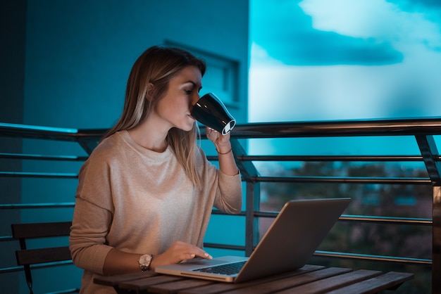 Caffè bevente della bella donna alla sera mentre per mezzo del computer portatile. Immagine ISO alta.